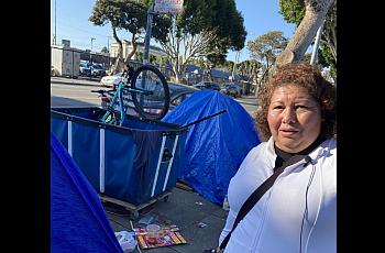 Person with microphone standing in front of blue tent like tarp on the sidewalk